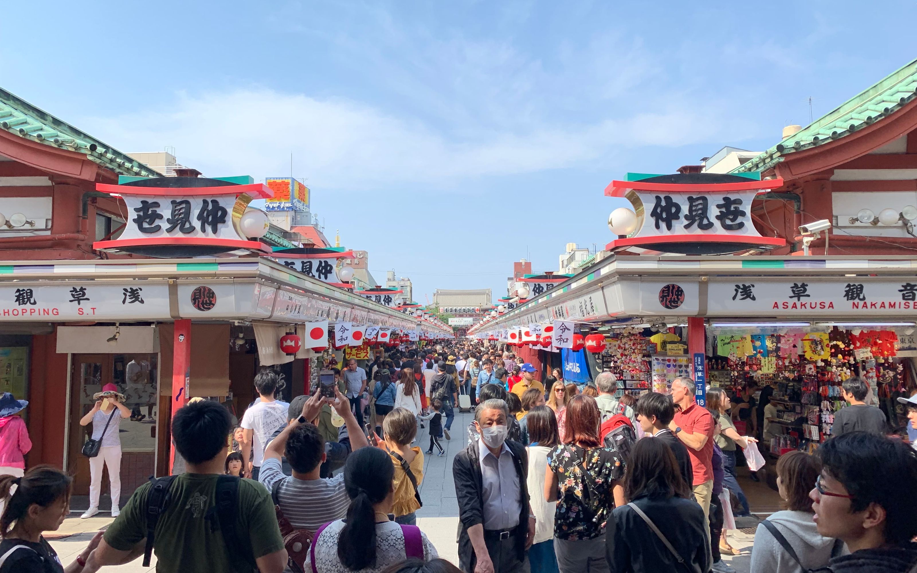 Crowded shopping street in Asakusa, Tokyo, lined with souvenir stalls and red lanterns leading up to a temple gate.