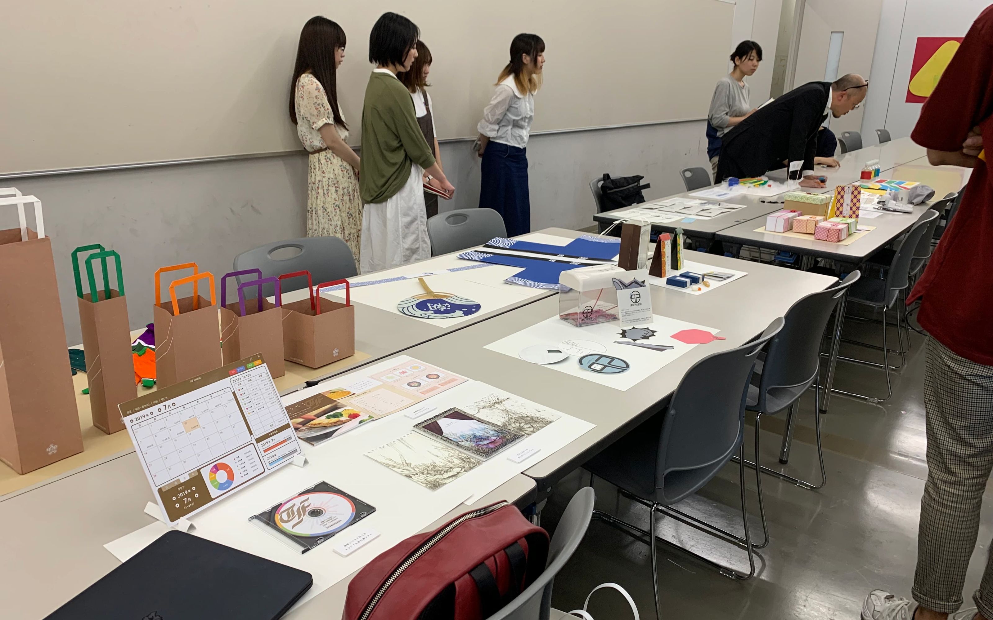 Course room with student work from a 'School Goods' assignment laid out on tables. Students designed packaging, gift bags and other prints.