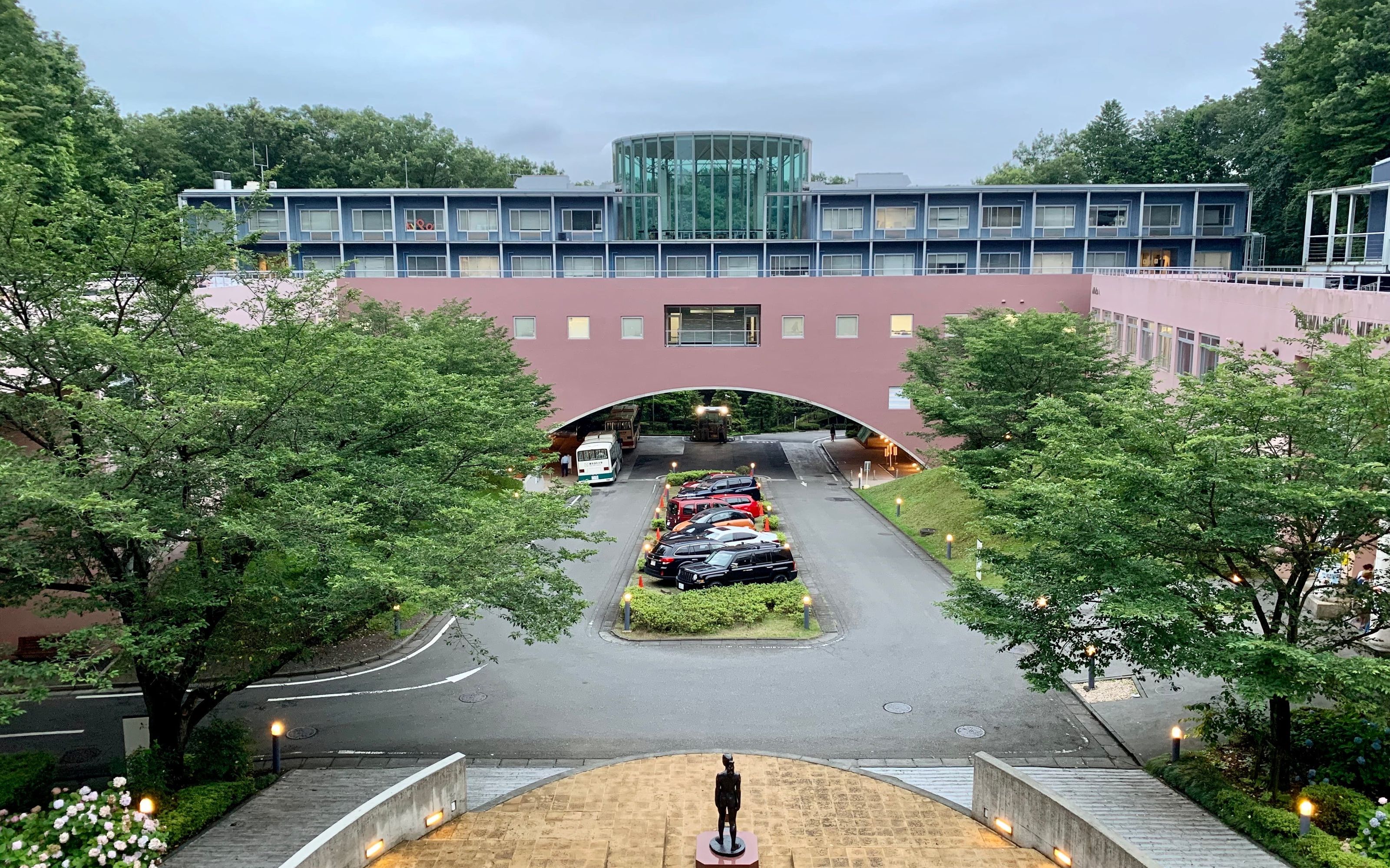 View of the campus entrance: a pink arched bridge building leading up to the professors' offices, with a small statue in the courtyard below.